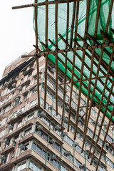 Low-angle perspective of traditional Hong Kong bamboo scaffolding framework with green mesh netting against a high-rise residential apartment facade. © Sergej