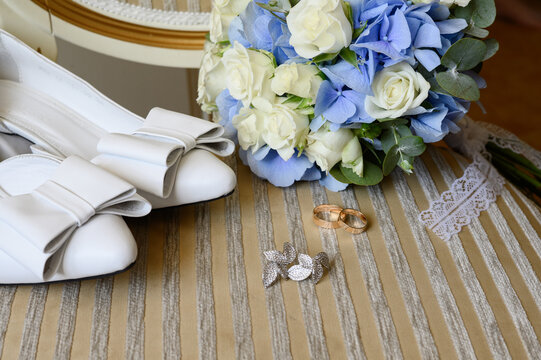 Elegant bride morning flat lay accessories with blue hydrangea bouquet and white bow shoes close up, modern wedding jewelry and luxury gold rings setup. Wedding preparation details.