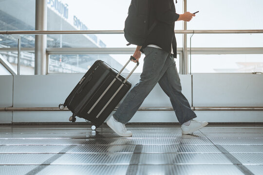 Traveler walking with rolling suitcase and smartphone through airport terminal, modern travel and journey concept