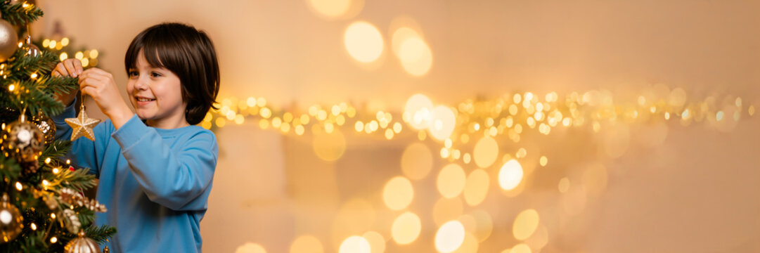 Smiling young boy decorating a Christmas tree with a gold star ornament. Wide festive banner with warm golden bokeh lights and copy space for text