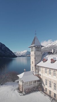 Dramatic aerial footage showcases a snow covered monastery nestled beside a calm lake and majestic mountains under a clear blue sky