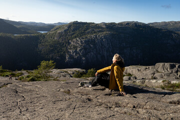 Fototapeta premium Individual wearing a yellow jacket sits on rocky terrain overlooking a vast mountainous landscape with trees and a lake in the distance under a clear blue sky