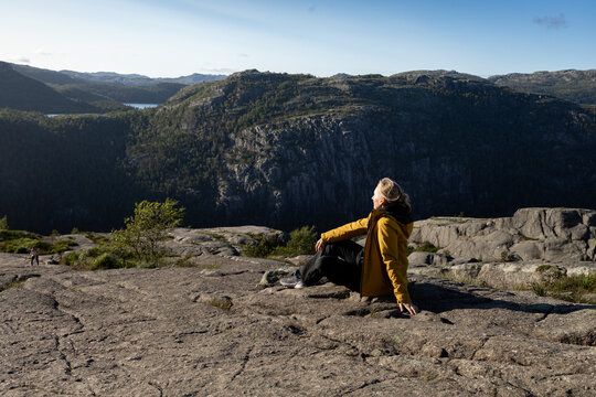 Individual wearing a yellow jacket sits on rocky terrain overlooking a vast mountainous landscape with trees and a lake in the distance under a clear blue sky