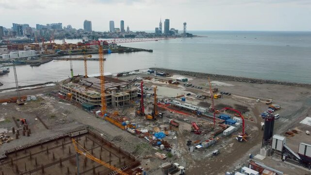 Extensive aerial photography showing a large-scale construction site on the coast of Batumi, with heavy machinery and foundation work against the backdrop of a modern city skyline and the sea in Georg
