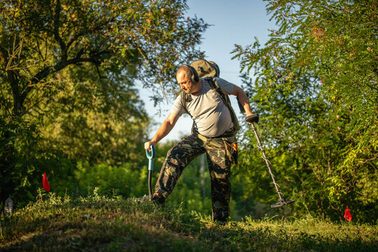 A treasure hunter stands in a green field using a metal detector. He wears camouflage pants and holds the detector in one hand. Red flags mark spots in the ground