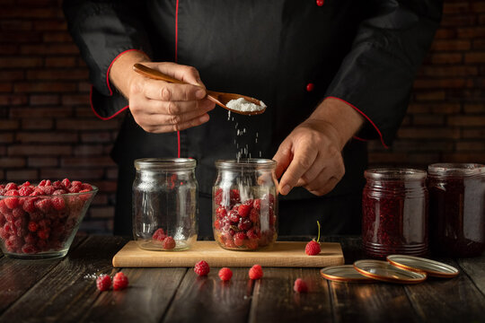A chef is adding sugar to a jar filled with fresh raspberries in a kitchen. Several jars and a bowl of raspberries are on the wooden table. It is daytime