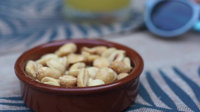 Macro shot of a small brown ceramic dish filled with salted peanuts, with a child's hand reaching in to pick up a peanut.