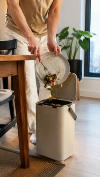 Person sorting compost waste from a plate into a modern compost bin in a bright indoor kitchen with plants and natural light streaming through a window