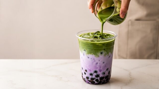 Ube matcha boba tea with purple ube milk, green matcha, black tapioca pearls and ice, finished by pouring matcha into a clear takeaway cup on a marble counter, styled for a cocktail aesthetic