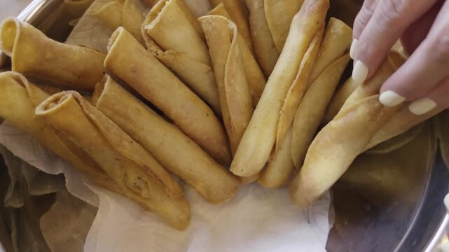 Top view of hand picking a crispy golden fried taquito from a metal serving bowl lined with paper
