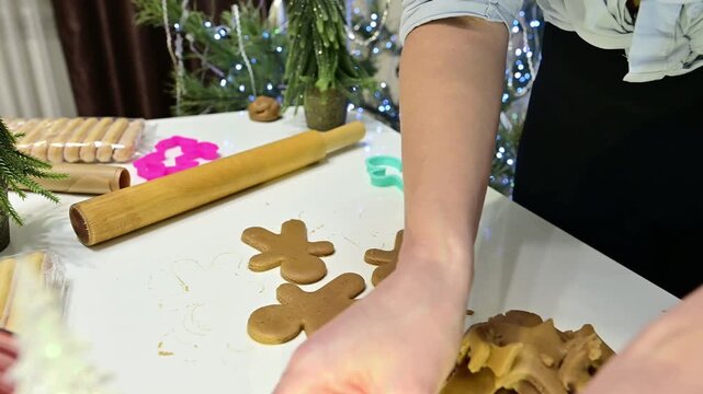 A girl prepares gingerbread dough at a table with a Christmas tree nearby. She rolls the dough and cuts out shapes of people for the holiday season.