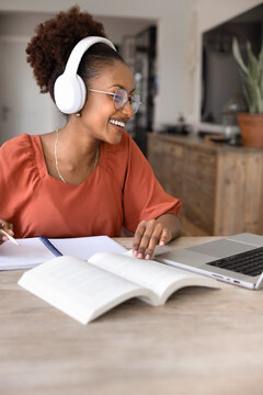 Happy African woman in headphones studying online sit at table with laptop, writes notes, making exercises, improve foreign language skills during videocall with teacher. Audio course, remote learning