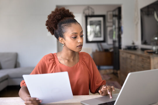 Focused African woman sits at desk, looking at laptop while holding printed document, engaged in remote work, financial planning or data review at home office. Digital workflow, accountancy, paperwork