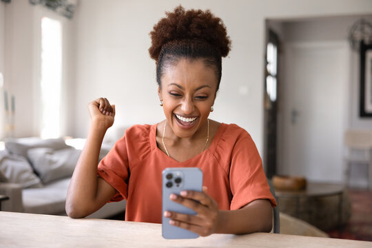 Excited young 25s African woman rejoices while reading great news in sms on her phone, raises her fist and smiles with joyful expression. Success celebration, positive results, lottery or betting win