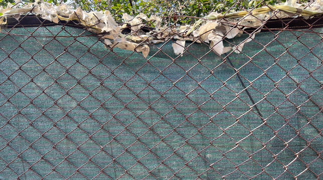 Close‑up of a rusted chain‑link fence in front of a green privacy screen, topped with torn fabric and tangled wires, creating a layered urban texture with geometric patterns and weathered detail.