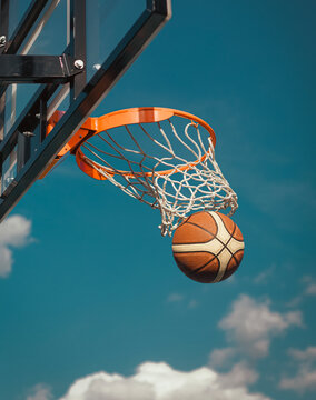 Basketball falling through the net of a hoop against a blue sky with clouds.