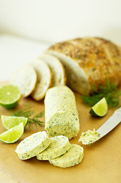 Fresh, homemade log of herbed butter with springs of dill, wedges of lime and a sliced loaf of bread slightly blurred in the background