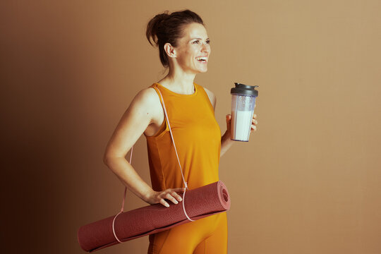 A smiling woman in an orange-yellow activewear outfit, with her hair in a bun, holds a yoga mat and a shaker bottle. Her cheerful pose suggests readiness for exercise and a healthy lifestyle.