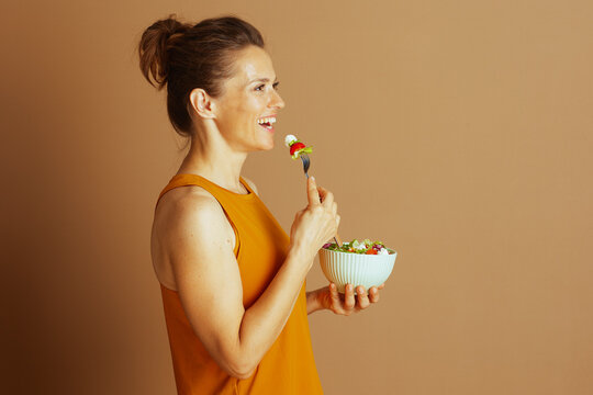A smiling woman in a mustard yellow top enjoys a fresh salad, bringing a forkful of tomato and mozzarella to her mouth, captured in a side profile.