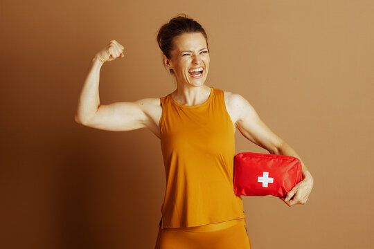 An energetic woman in orange activewear flexes her bicep, holding a first-aid kit. Her confident smile conveys strength, fitness, and preparedness.