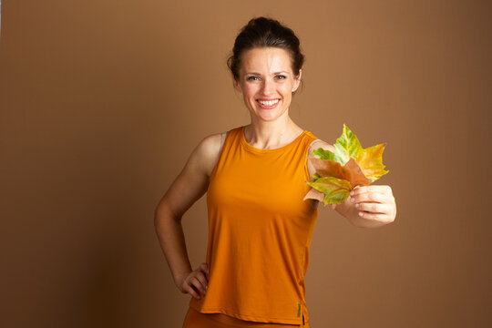A smiling middle-aged woman in mustard yellow activewear holds a cluster of autumn leaves towards the viewer, conveying a cheerful and healthy seasonal atmosphere.