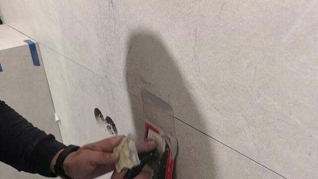 A worker grouts tiles with epoxy grout. A man is repairing damaged wall tiles in a house. He uses tools to remove the old material and prepare the surface for new tiles. The work is being done near 