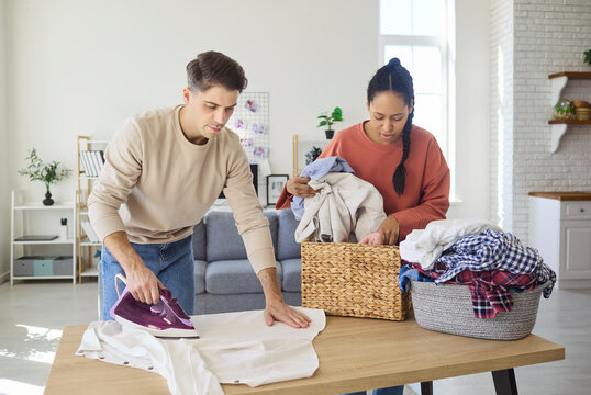 Couple ironing laundry at home with teamwork. Man and woman share chores in a bright apartment, sorting clothes and pressing shirts on a table. Clear concept of modern shared housework.