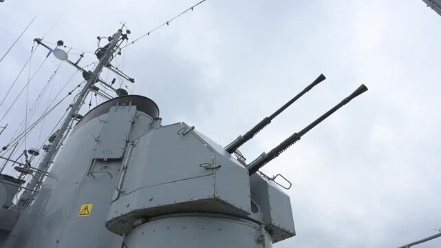 Decommissioned dual-barrel naval cannon on a warship deck with military mast background.
