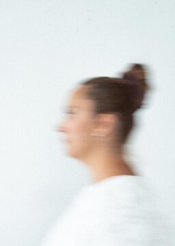 A blurred side-profile portrait of a young woman with her hair tied up in a bun, looking to the left against a clean, light background. Soft focus and minimal composition create a modern, natural aest