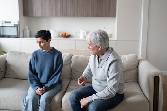 Angry elderly man shouting and blaming wife while she sits silently listening, senior couple facing domestic conflict at home. Narcissistic outbursts, emotional abuse, manipulative control. 