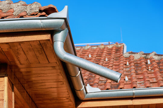 Bright sunny close-up of a new galvanized steel gutter and downspout elbow on a wooden eave, terracotta roof tiles beyond.