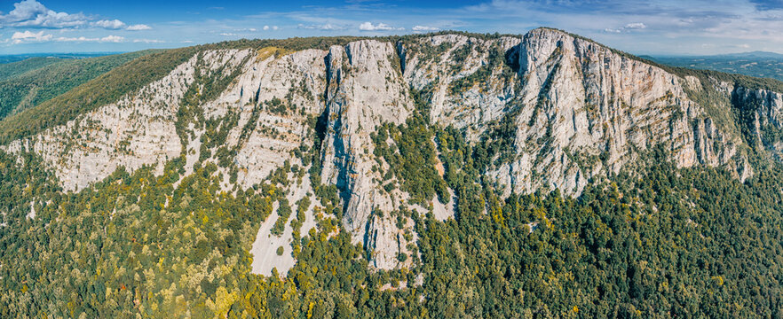 Aerial panoramic view of dramatic limestone cliffs bathed in warm autumn light, surrounded by dense forested slopes under a bright blue sky. Nature concept.