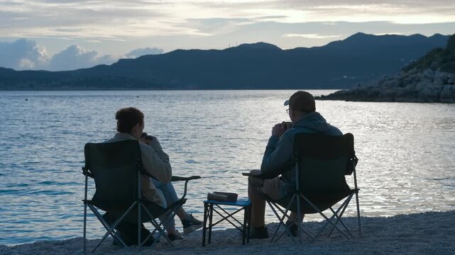 Couple relaxing on camping chair, enjoying tranquil sunset view. Couple relaxing on camping chair, sitting side by side while watching sunset and distant mountainous landscape with soft evening light