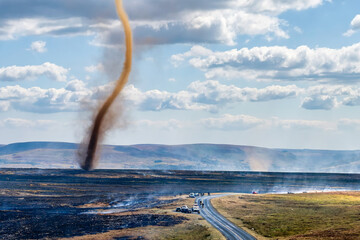 Tall smoke whirl spinning above scorched moorland terrain, emergency vehicles parked by rural road...