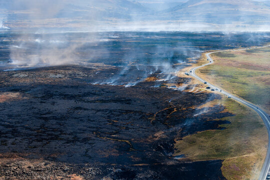 High-angle view of charred landscape after moorland wildfire, smoke drifting over burnt ground alongside asphalt road under hazy daytime sky