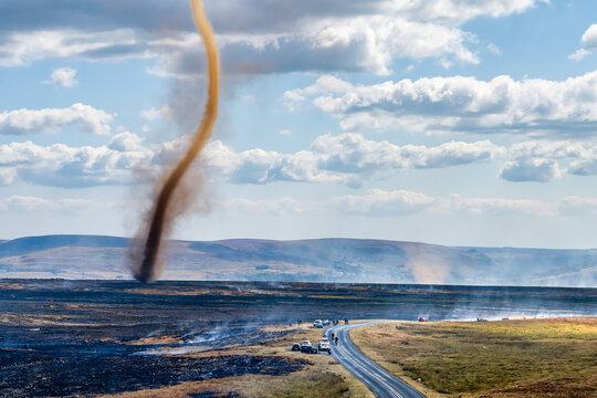 Tall smoke whirl spinning above scorched moorland terrain, emergency vehicles parked by rural road under cumulus cloud sky, Brecon Beacons area