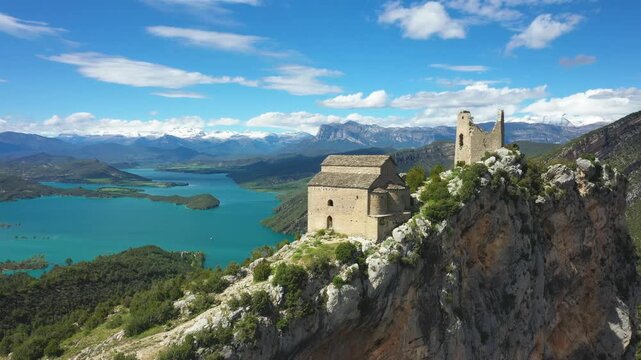 Stunning aerial footage of Samitier Castle perched dramatically on a rocky cliff above a turquoise lake, surrounded by lush green hills and distant mountain ranges.