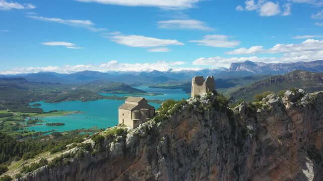 Drone footage of the medieval Samitier Castle and chapel perched on a dramatic rocky ridge, overlooking a turquoise reservoir and green valleys in the Pyrenees foothills near Huesca, Aragon, Spain.