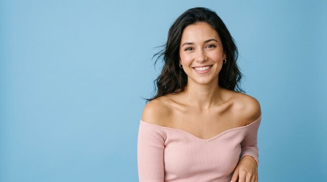 Smiling young brunette woman in soft pink off-shoulder top posing against clean sky blue studio background with natural makeup and wavy hair
