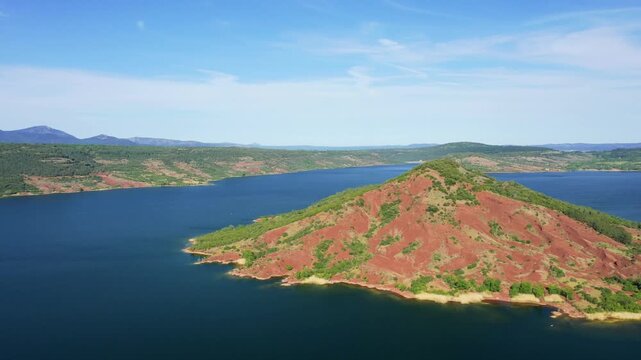 Aerial footage showcasing the striking red hills and deep blue waters of Lac de Salagou, surrounded by green fields and rolling hills in the Herault department, Occitanie region, southern France.