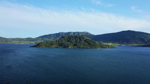 A wide aerial shot of Lac de Salagou in Herault, Occitanie, France, featuring a prominent green island surrounded by deep blue water.