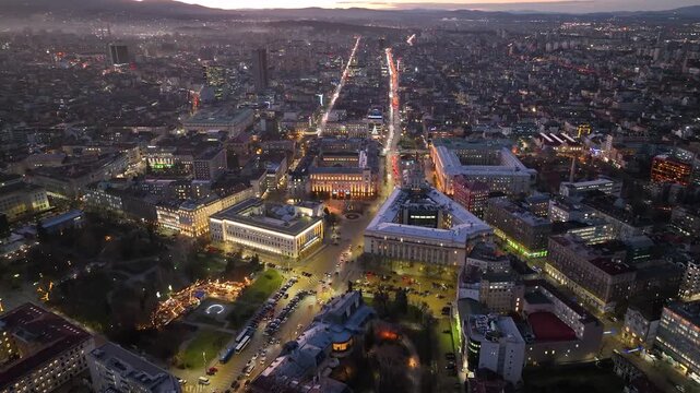 Aerial footage of Sofia, Bulgaria at dusk, showing the illuminated city center, busy boulevards, historic buildings, urban skyline and Vitosha Mountain in the background.