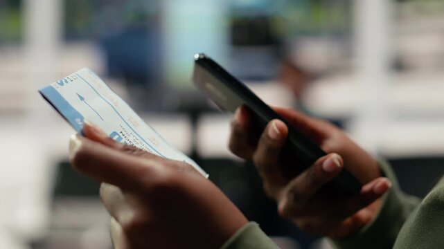 Traveler holds a bus ticket in a shuttle terminal while scrolling on a phone, checking the boarding pass and route itinerary before departure.