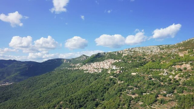 Aerial footage showcasing the picturesque village of Baunei nestled on a lush green mountainside under a bright blue sky with scattered clouds.