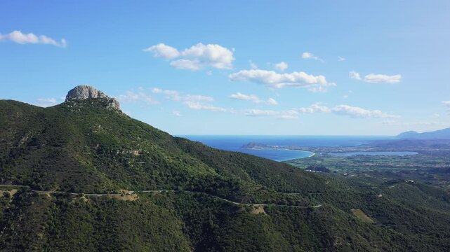 Drone footage showing a rugged green mountain landscape with a prominent rocky peak and winding road, overlooking the blue Mediterranean Sea and sweeping coastline between Baunei.