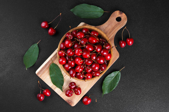 Fresh ripe sweet cherries in wooden bowl on rustic cutting board with green leaves and linen cloth on black textured background. Healthy summer fruit, red berries, fresh seasonal harvest, healthy food