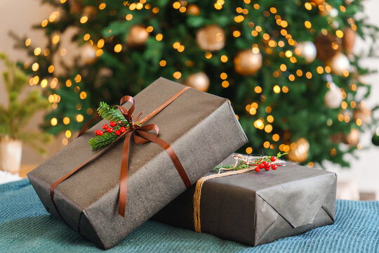 Two holiday gift boxes decorated with brown ribbon, evergreen sprig and red holly berries in front of glowing Christmas tree with warm golden bokeh lights background. Celebration, winter holidays mood