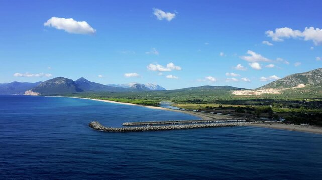 Aerial footage of the entrance to the marina at Orosei, Sardinia, Italy. Rugged mountains and a partly cloudy sky complete the scenic coastal panorama. No people are visible in the scene.