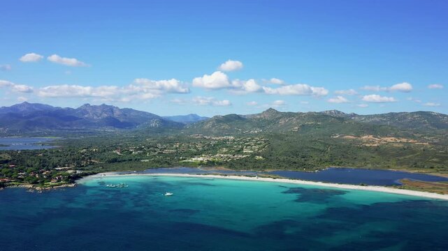 A high-altitude aerial shot of Spiaggia Lu Impostu beach near San Teodoro in Sardinia, Italy. Several boats are anchored in the clear shallows, and the sky is mostly clear with scattered clouds.