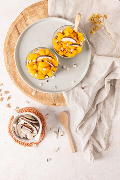 Two glass bowls filled with fresh diced mango and coconut slices sit on a ceramic plate and wooden tray served with a side of coconut flakes and a linen cloth in a bright aesthetic setting